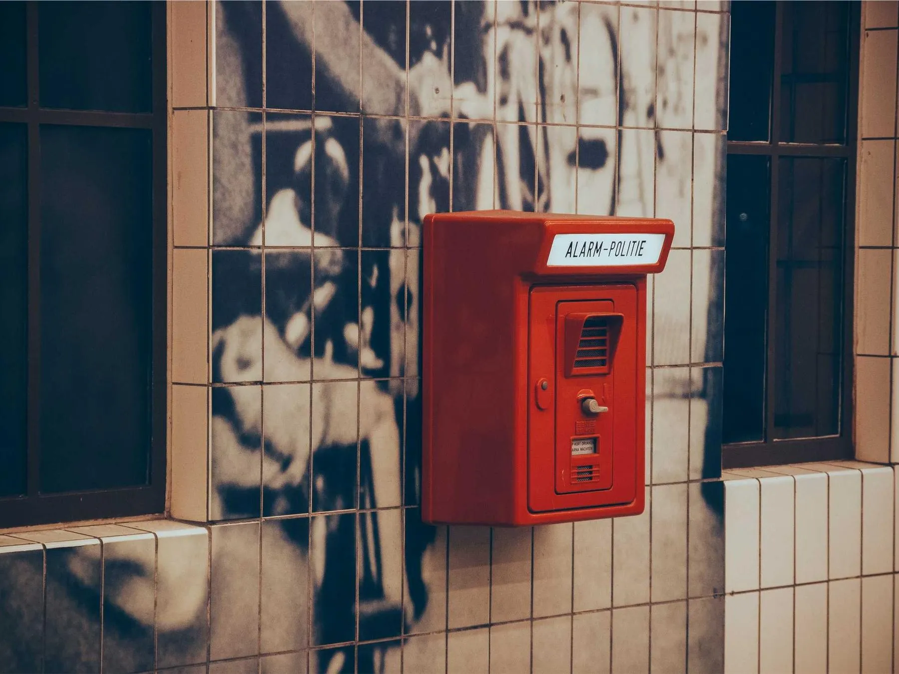 A red emergency alarm box mounted on a tiled wall beside a black-and-white mural